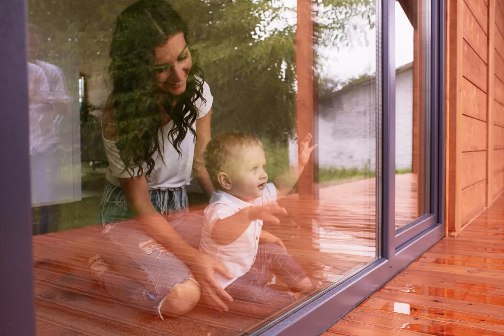Mãe e filho pequenos sentados próximos a uma porta de vidro, observando o lado externo em um ambiente acolhedor.
