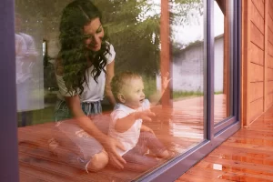 Mãe e filho pequenos sentados próximos a uma porta de vidro, observando o lado externo em um ambiente acolhedor.